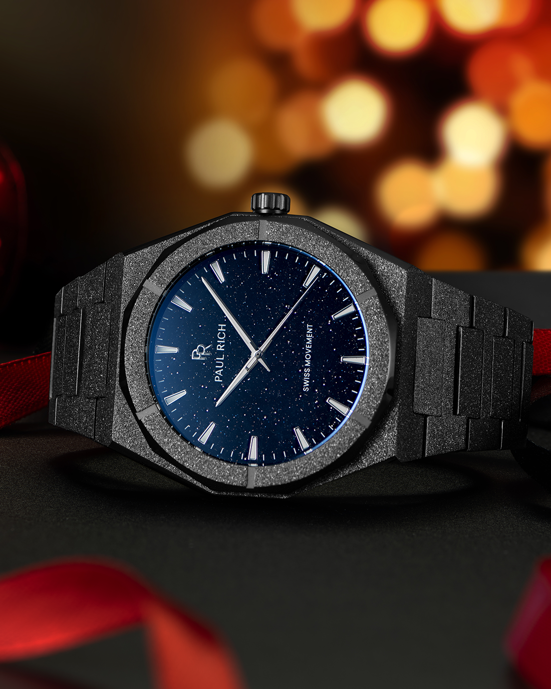 Black frosted Paul Rich watch with blue aventurine dial on black surface, surrounded by red ribbon and warm holiday lights in background.