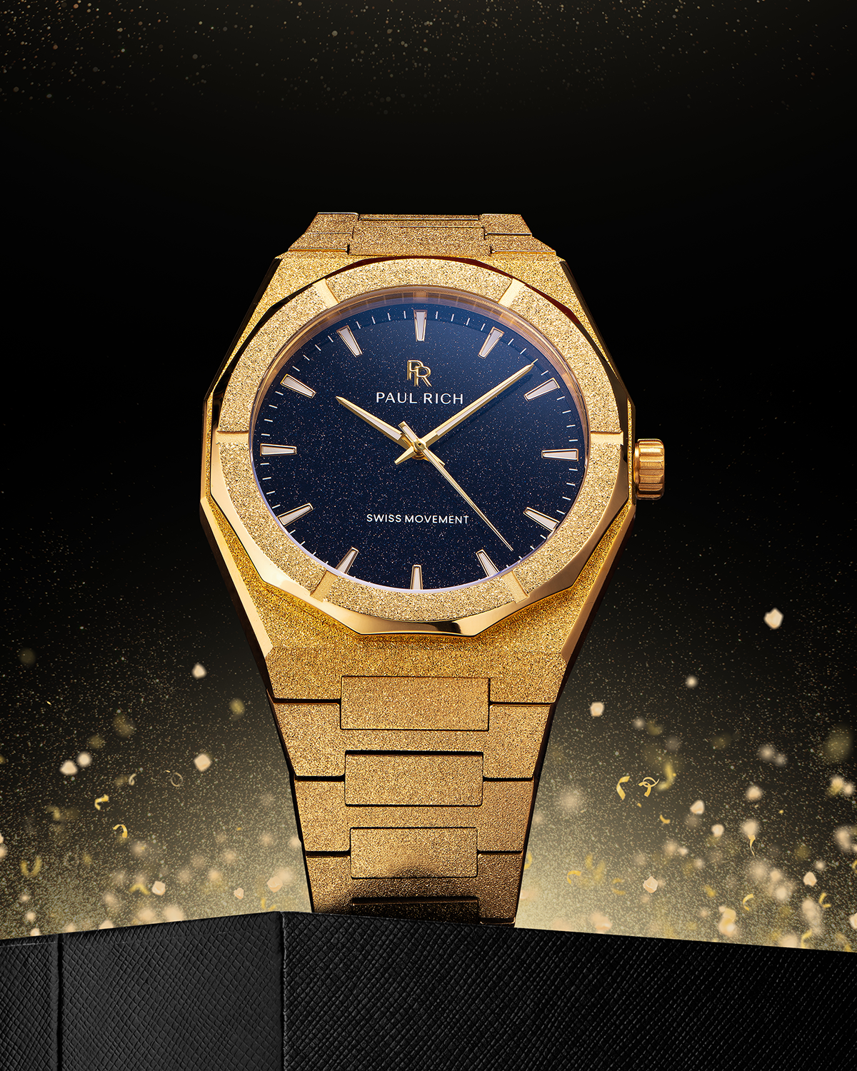 Gold frosted Paul Rich watch with blue aventurine dial displayed upright on textured black surface, with golden sparkles rising behind.