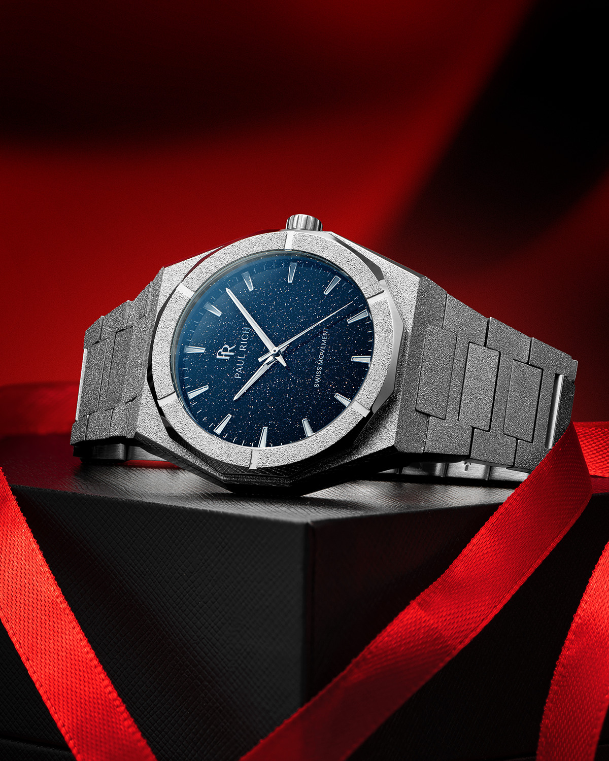 Silver frosted Paul Rich watch with a deep blue aventurine dial, placed on a black gift box with red ribbon on red-black background.