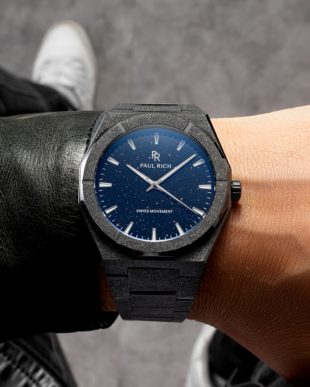 Black frosted Paul Rich watch with blue aventurine dial and silver markers, worn on wrist with black leather jacket and sneakers.