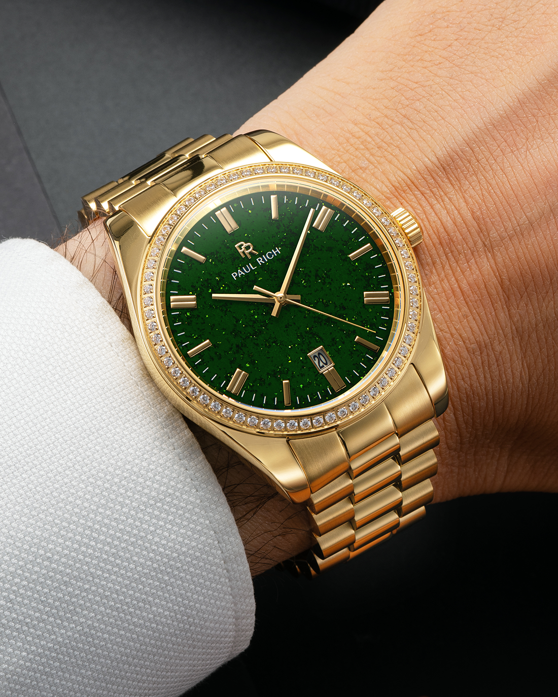 Gold-tone Paul Rich watch with green aventurine dial, diamond-set bezel, and gold link bracelet, worn on wrist with a white textured dress shirt cuff.