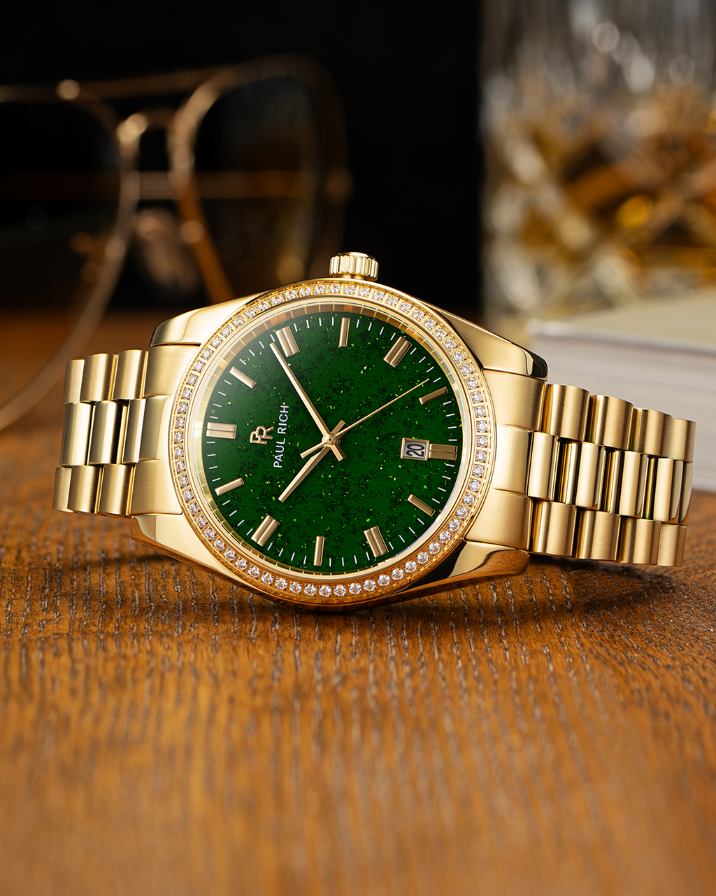 Gold-tone Paul Rich watch with green aventurine dial, diamond-set bezel, and gold link bracelet, displayed on a wooden surface with a pair of sunglasses and a glass tumbler in the background.