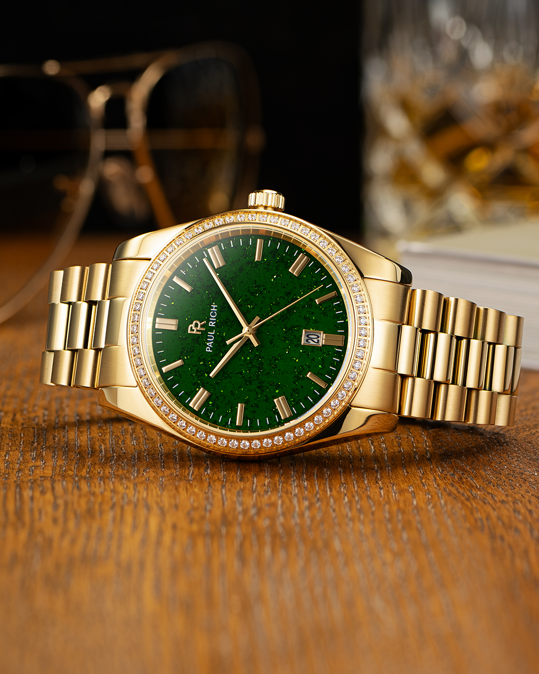 Gold-tone Paul Rich watch with green aventurine dial, diamond-set bezel, and gold link bracelet, displayed on a wooden surface with a pair of sunglasses and a glass tumbler in the background.