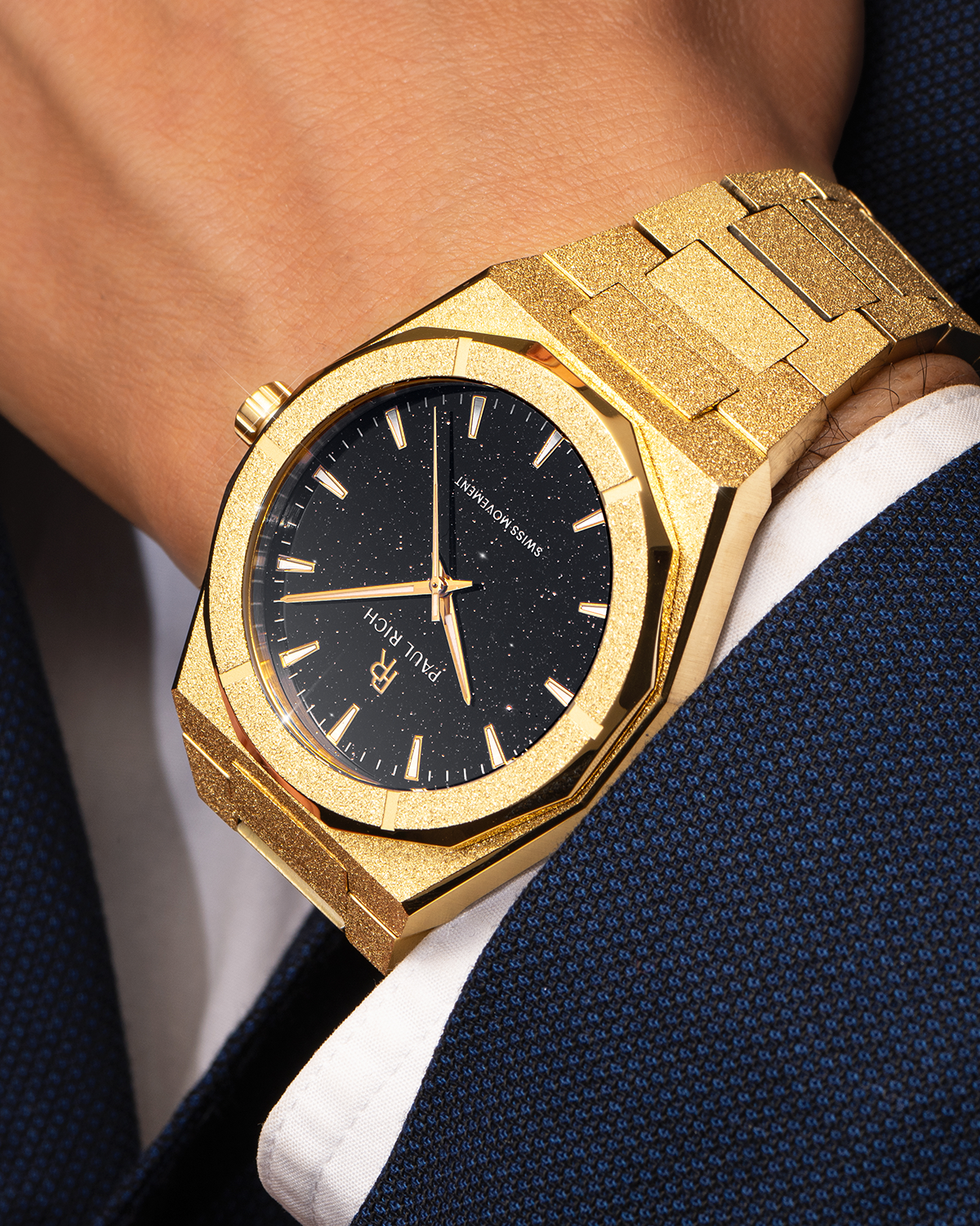 Gold Paul Rich watch with black aventurine dial and frosted finish, worn on wrist with a dark blue textured suit and white cuff.