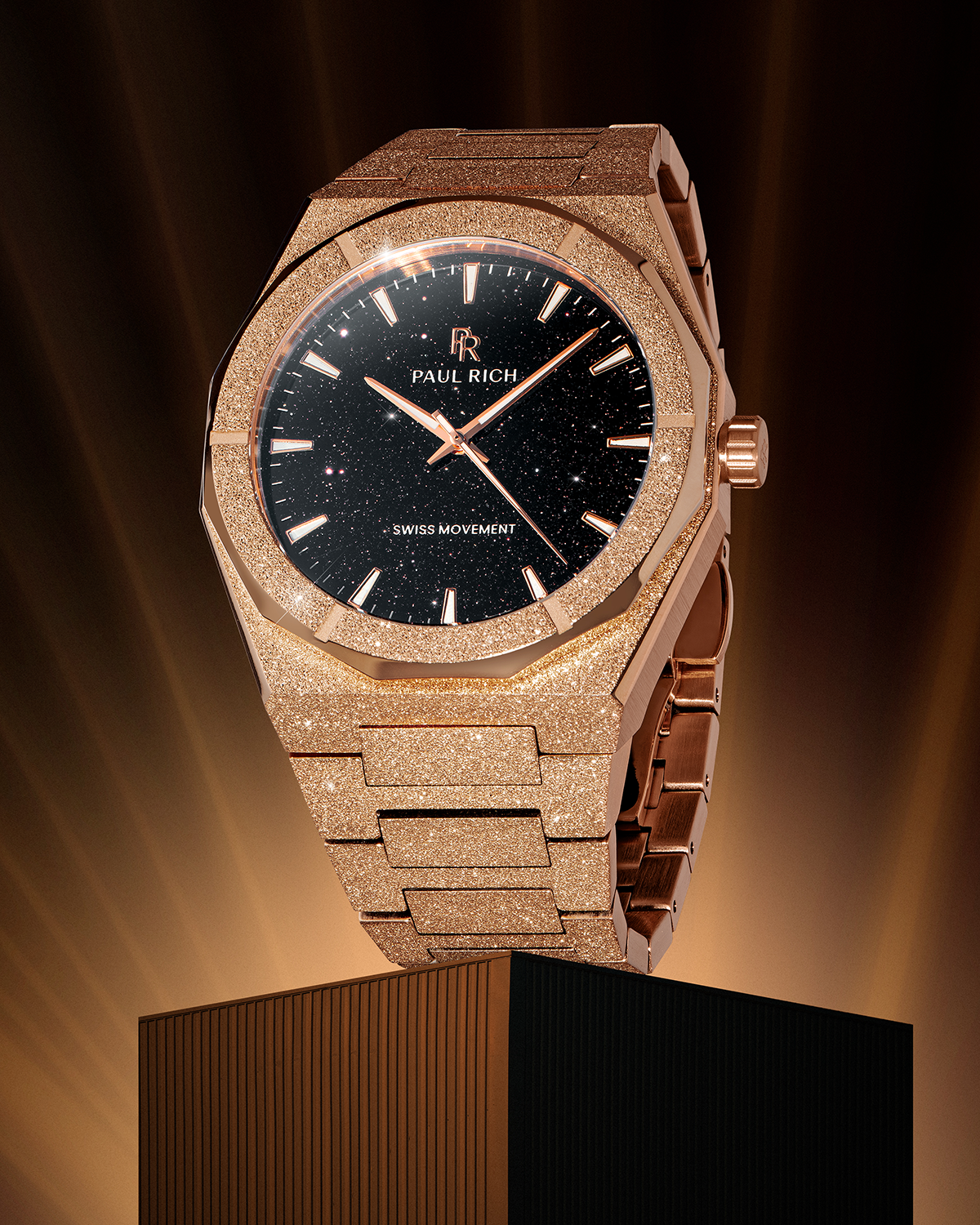 Rose gold Paul Rich watch with fully frosted case and bracelet, black aventurine dial, rose gold markers and hands, and labeled “Swiss Movement,” displayed upright on a pedestal with dramatic golden lighting.