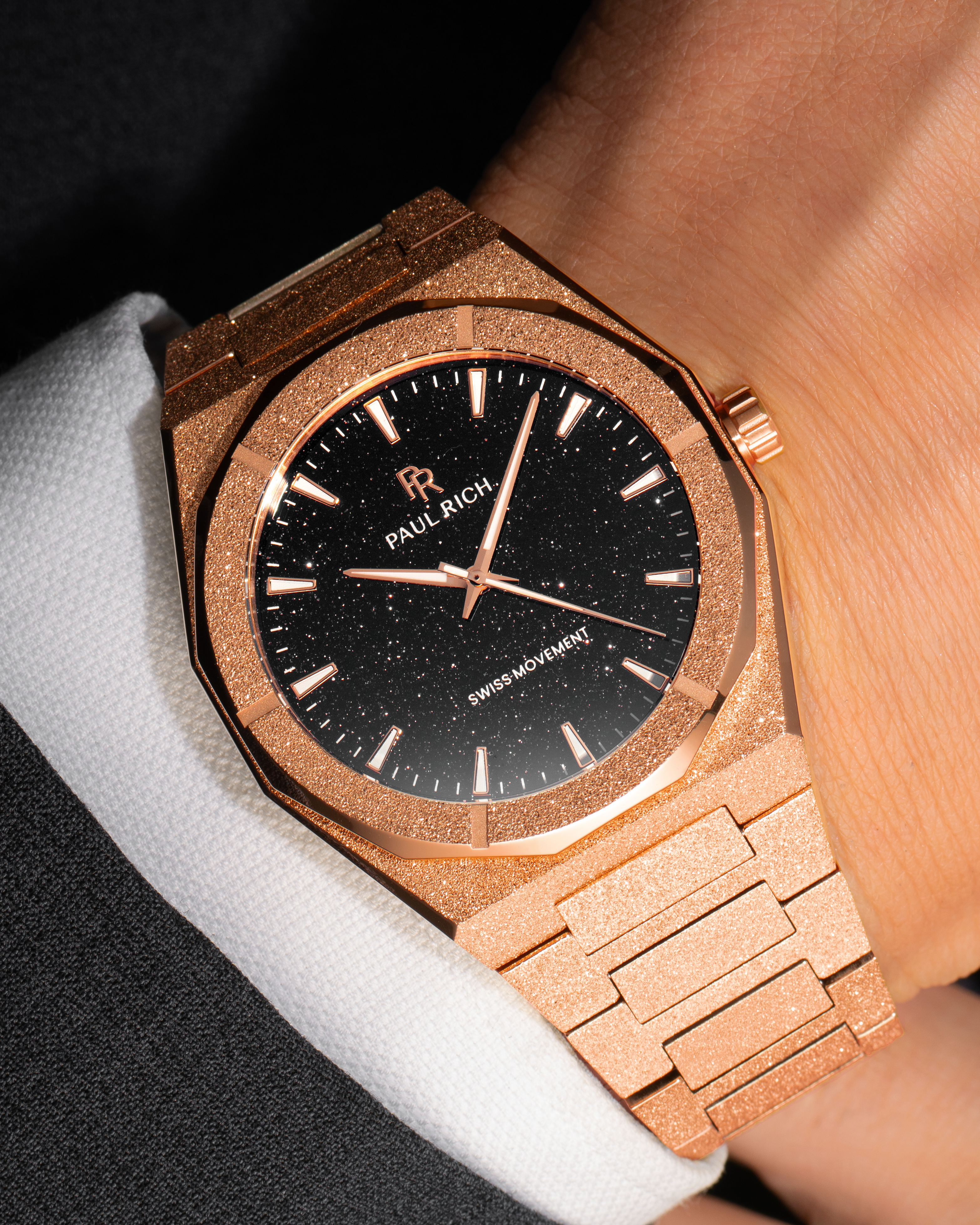 Rose gold Paul Rich watch with fully frosted case and bracelet, blue aventurine dial, rose gold markers and hands, worn on wrist with a white cuff and black suit.