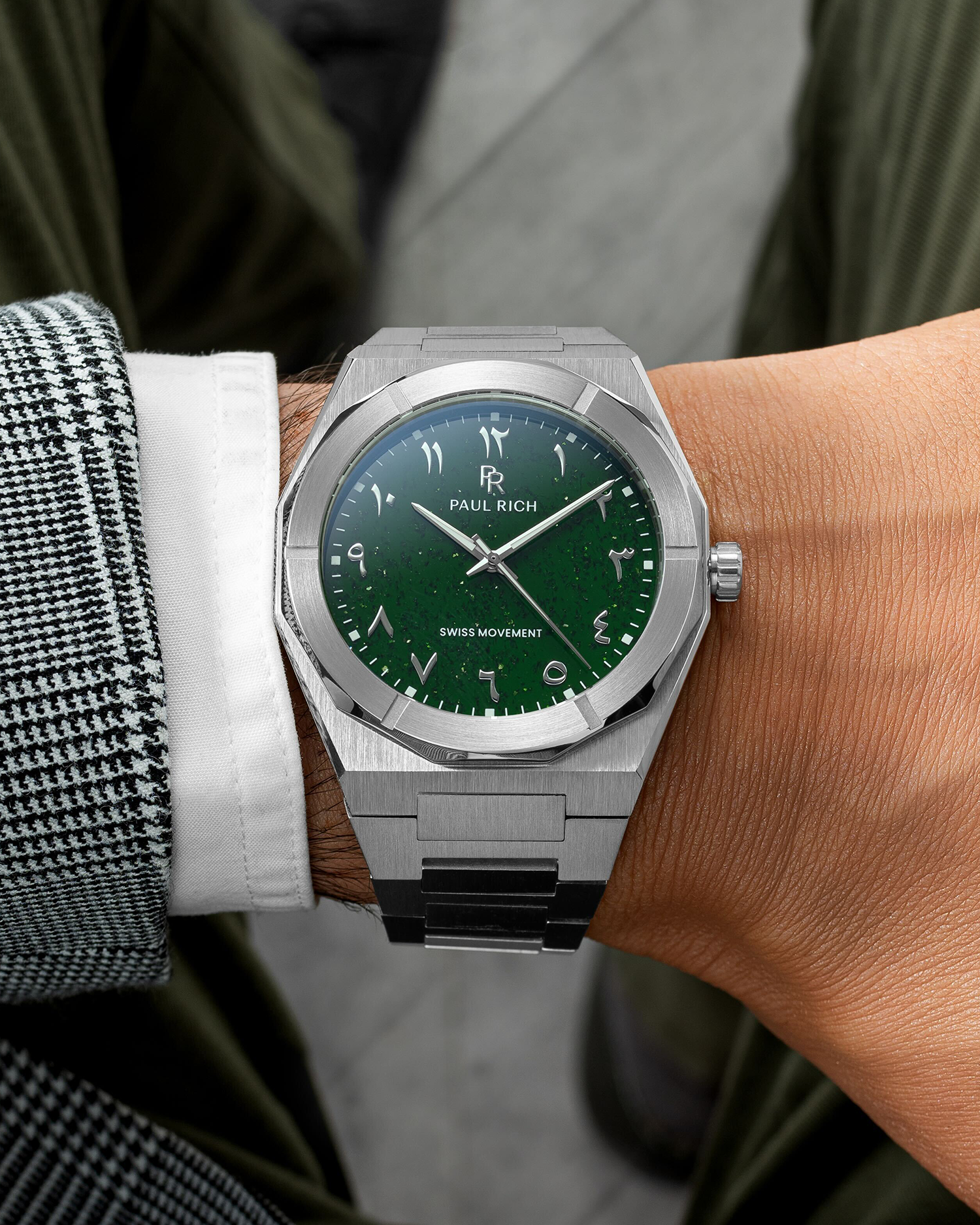 Paul Rich watch with brushed silver case and green dial featuring Arabic numerals, worn with a white cuff and houndstooth jacket.