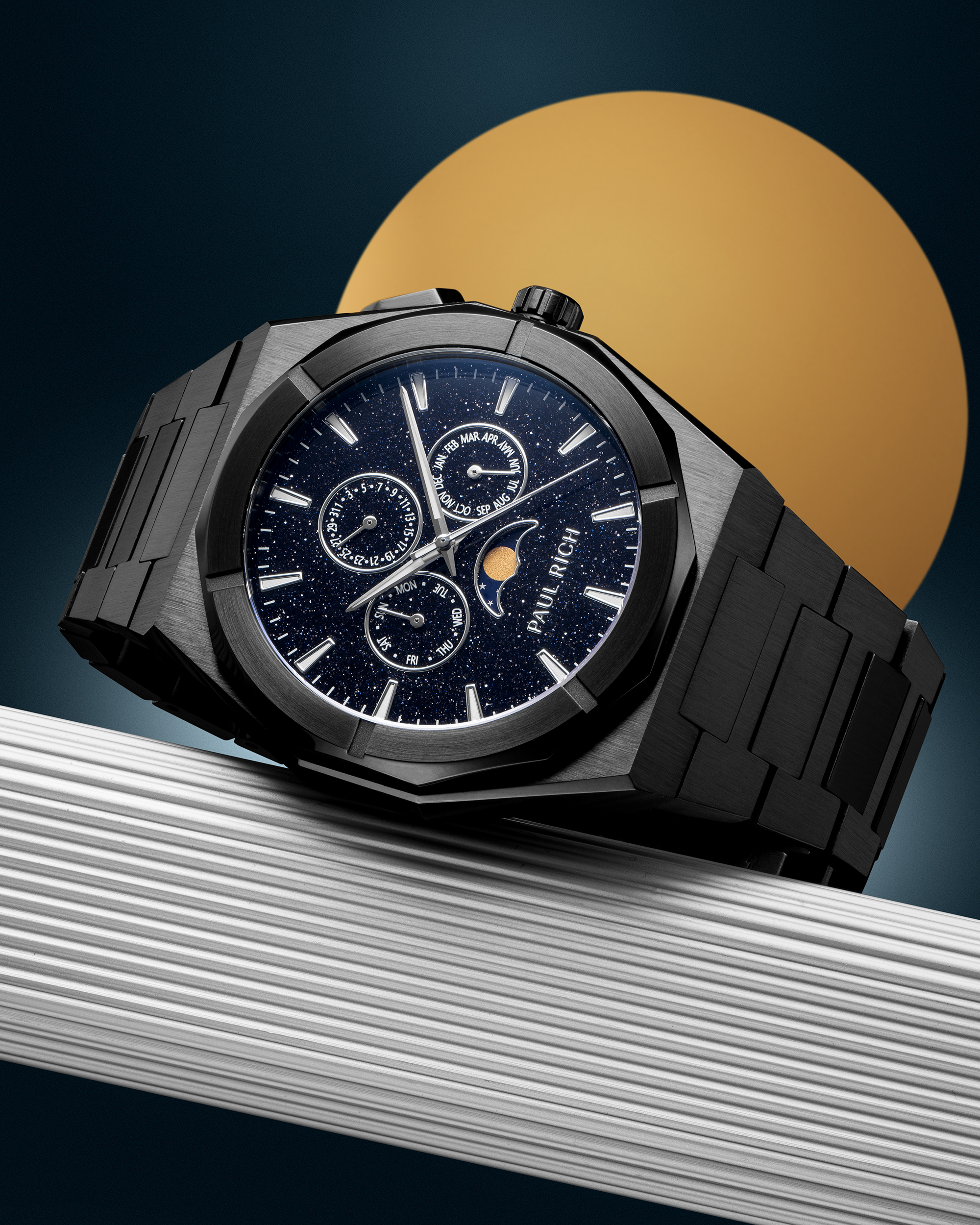 Black stainless steel watch with blue aventurine dial, moonphase, and subdials, displayed on a ribbed silver surface against a gold circle backdrop.