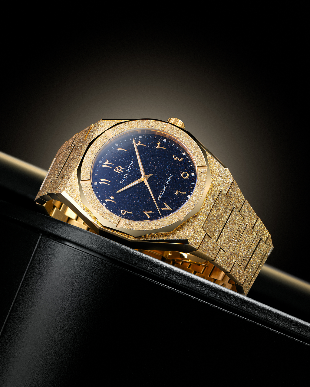 Frosted gold watch with blue aventurine dial and Arabic numerals, displayed on a black leather surface with a spotlight gradient background.